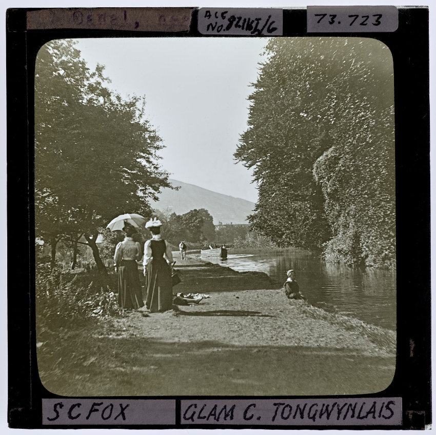 Two ladies walking along tow path. A small child sat on the edge of the canal. A barge being pulled by a horse.